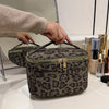 A dark brown leopard print cosmetic bag is being held by a hand on a white surface.