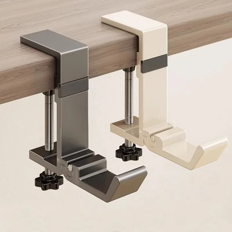 A close-up view of a foldable aluminum desk hook in gray and cream colors mounted on a wooden desk surface.