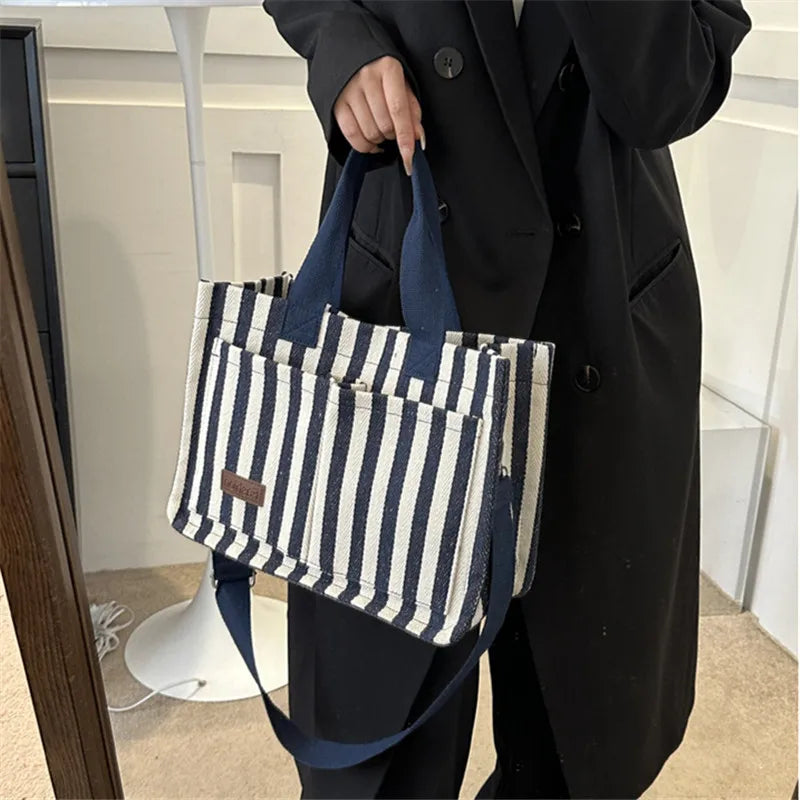 A person holds a striped canvas tote bag with dark blue straps and inner pockets against a white background.