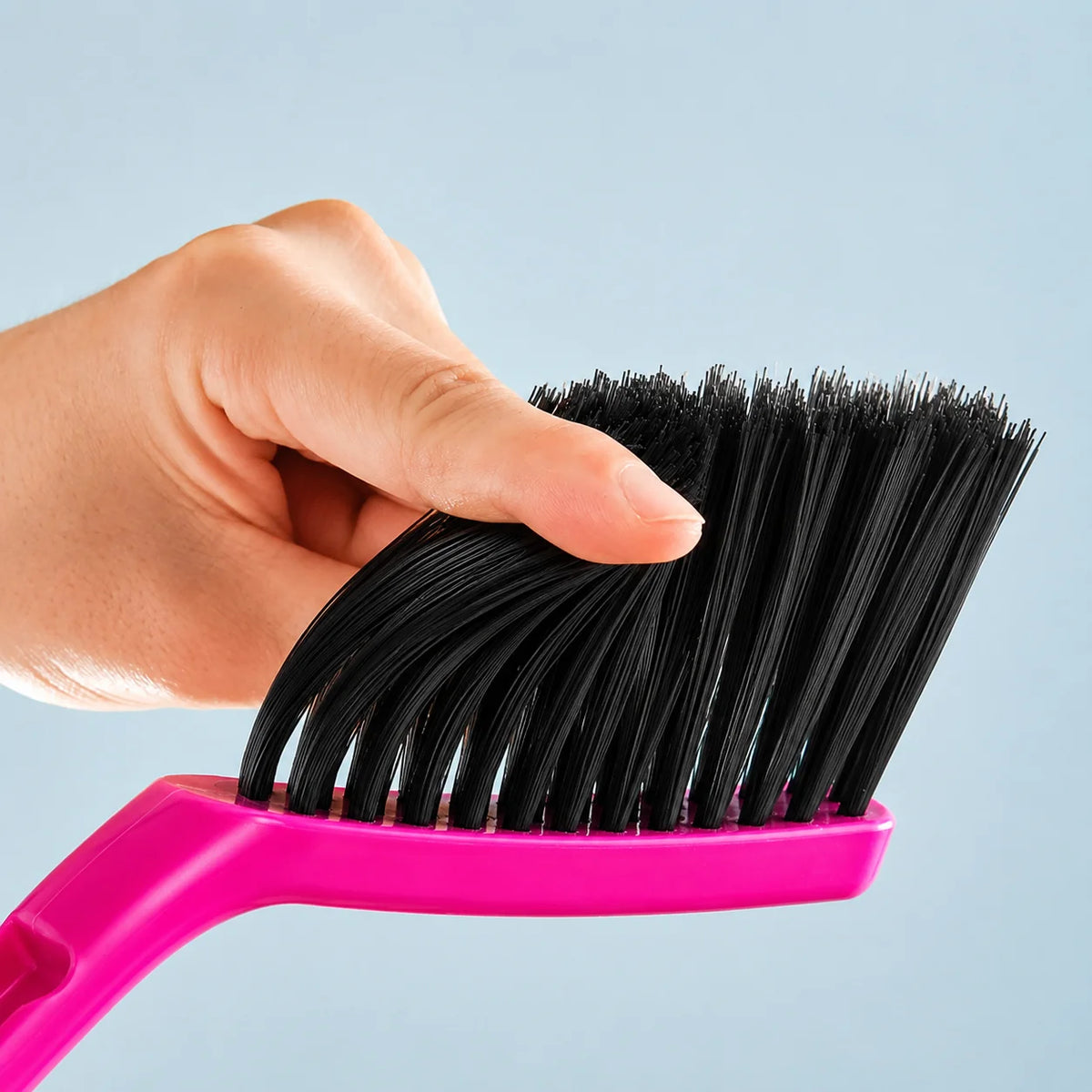 A hand holds a pink compact handheld dustpan and brush set with black bristles against a light blue background.