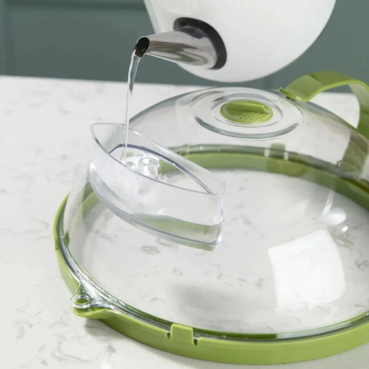 A clear microwave splatter cover with a steamer nozzle and green handle is shown pouring liquid onto a plate.