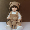 A lifelike silicone baby doll wearing a knitted hat and overalls with a teddy bear, sitting on a white surface.