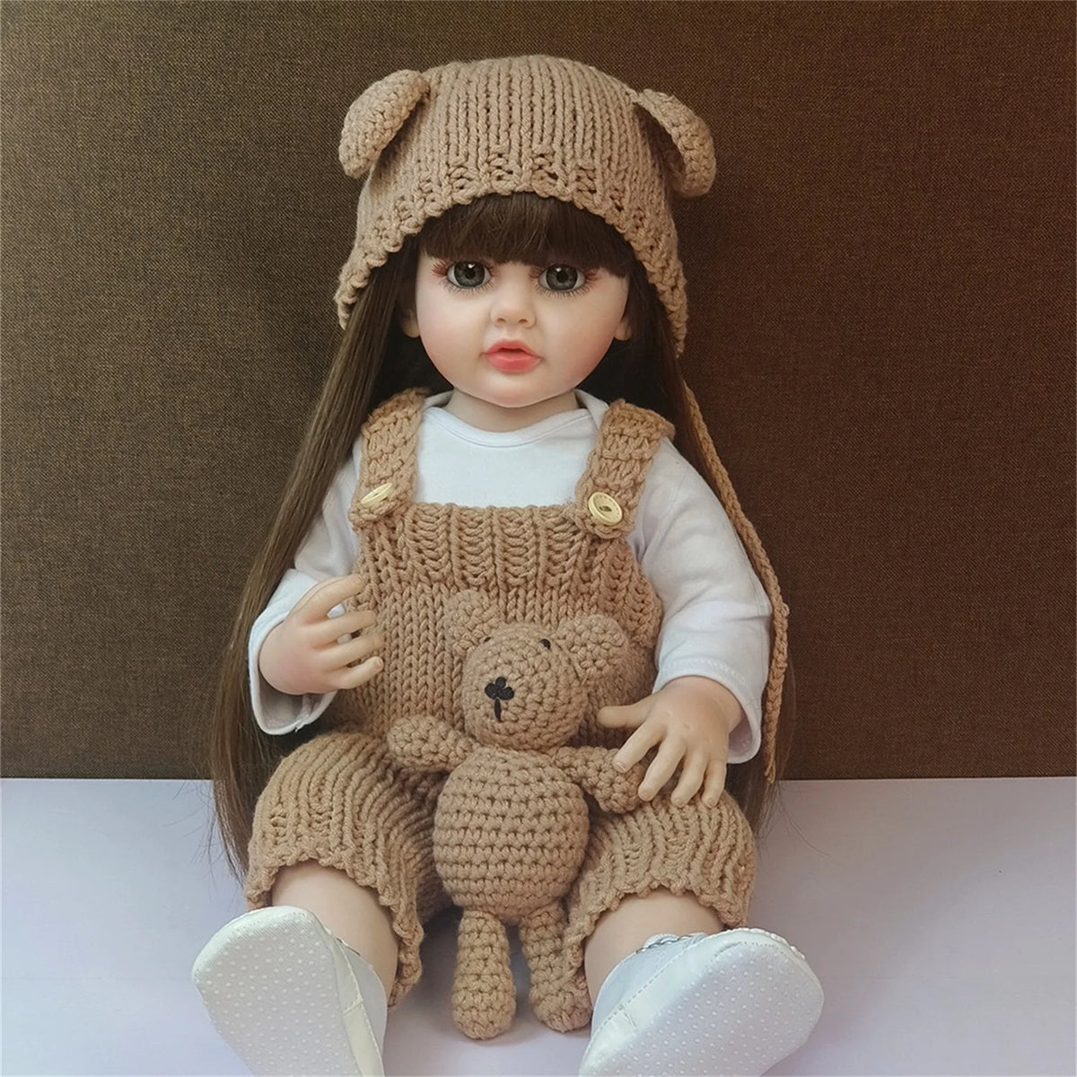 A lifelike silicone baby doll wearing a knitted hat and overalls with a teddy bear, sitting on a white surface.