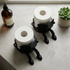 A black, human-shaped freestanding toilet paper holder stands next to a roll of white toilet paper and an oil bottle on a lig