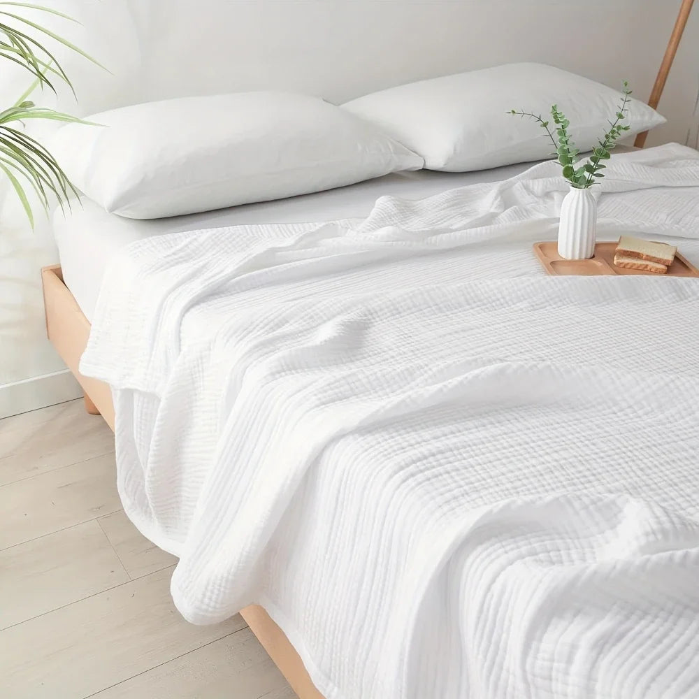 Eine weiße gesteppte Decke ist über einem Bett drapiert, mit einem weißen Kissen und einer Vase mit Grünzeug sichtbar.