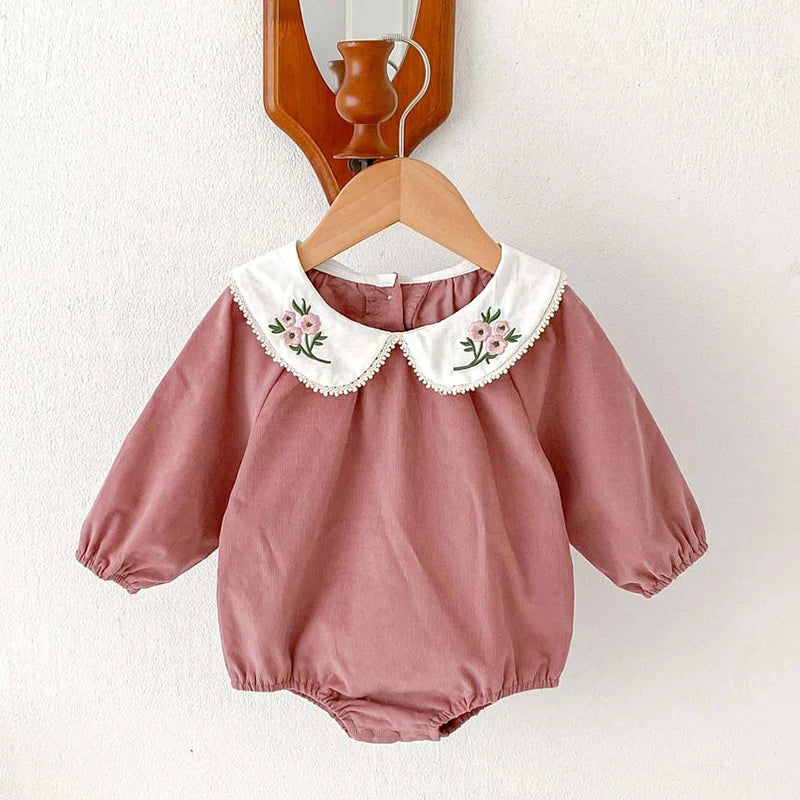 A mauve cotton romper for a baby girl featuring white floral lace trim, displayed on a wooden hanger.