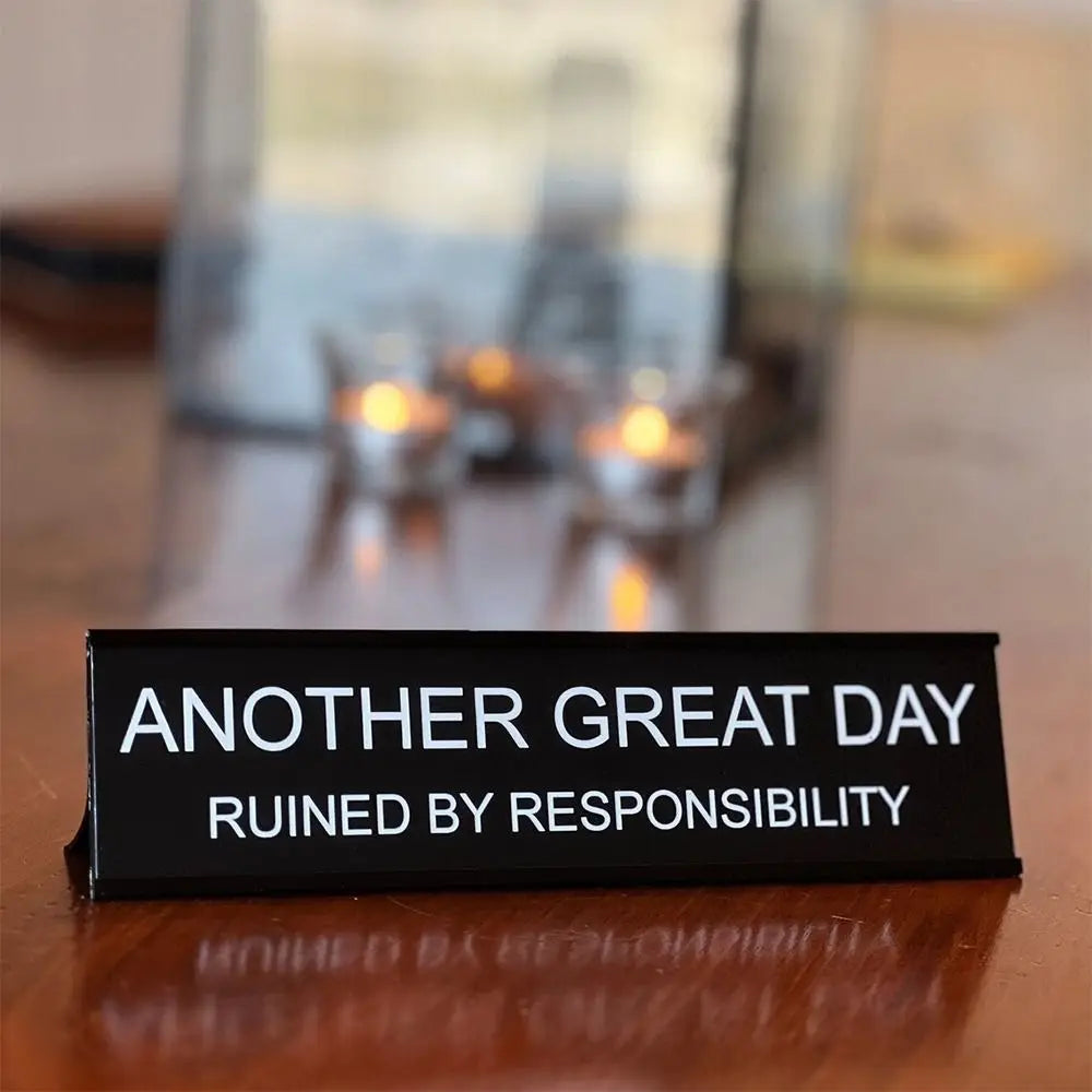 A black desk sign with white text reads Another Great Day Ruined by Responsibility