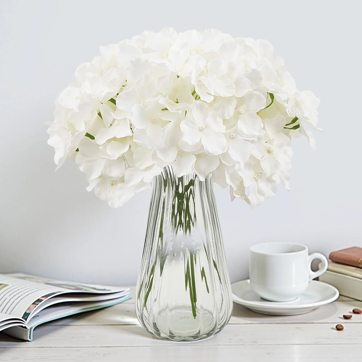 A clear glass vase filled with white hydrangea flowers sits on a white wooden surface.