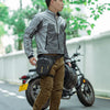 A young man wearing a gray jacket and brown pants stands next to a black motorcycle with a large waterproof hip pack.