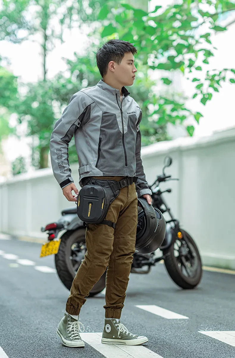 A young man wearing a gray jacket and brown pants stands next to a black motorcycle with a large waterproof hip pack.