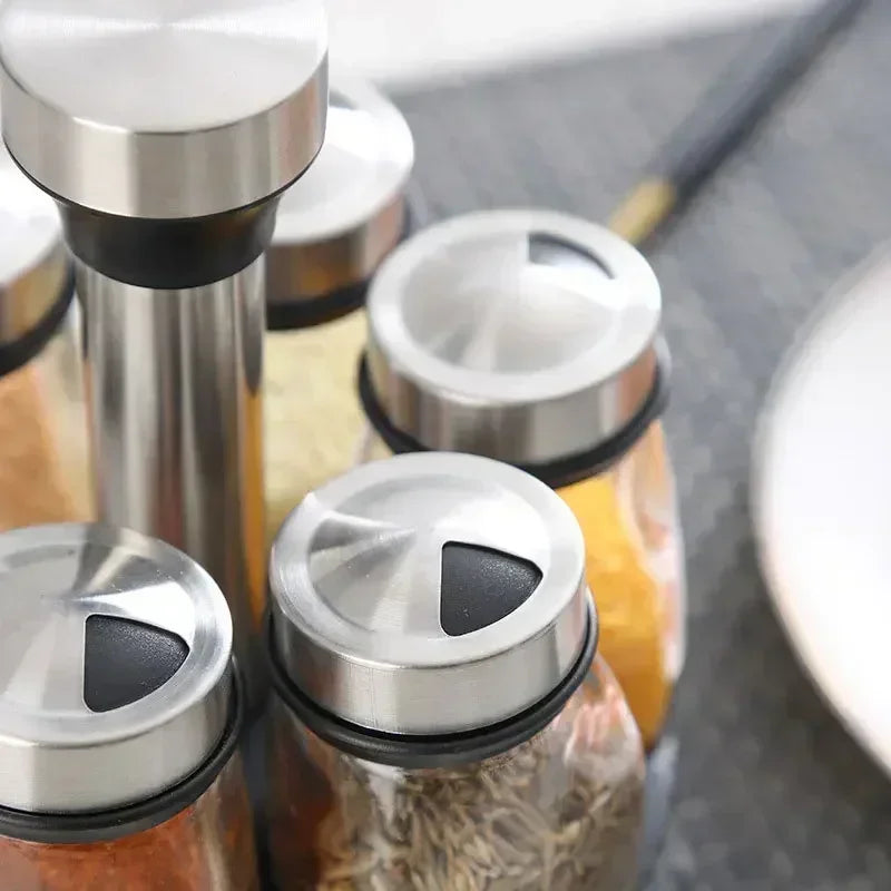 A close-up view of a stainless steel rotating glass spice jar set with a rack on a dark surface.
