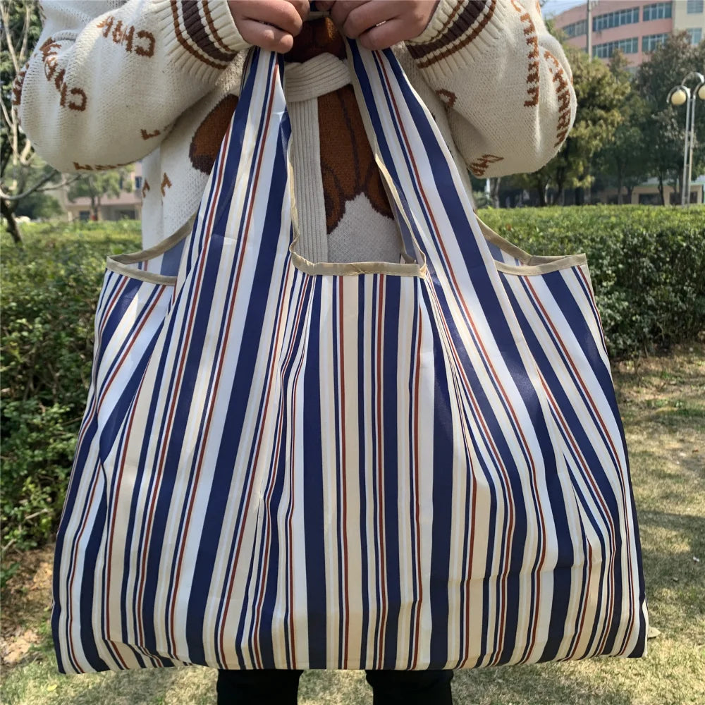 A person holds a striped blue and white tote bag made of Oxford cloth fabric outdoors.