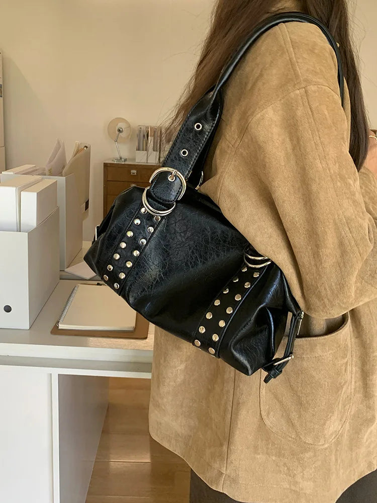A woman wearing a tan jacket carries a black soft PU leather shoulder bag with silver rivet accents.