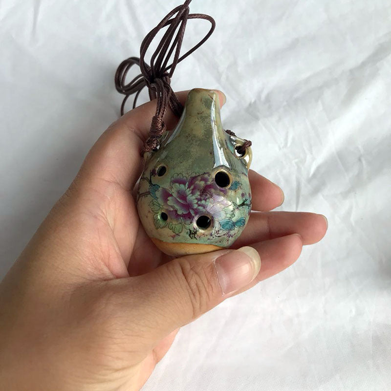 A hand holds a handcrafted ceramic pendant ocarina with six holes and a leather cord.