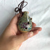 Handcrafted ceramic pendant ocarina with six holes held in a person's hand.