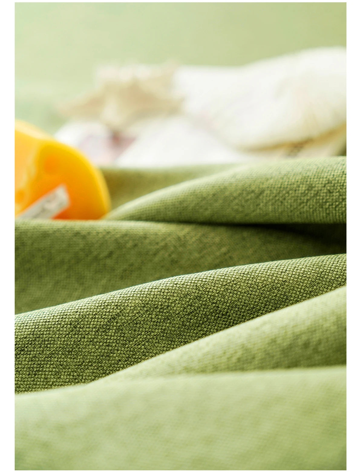 A close-up of a textured olive green farmhouse tablecloth with white cotton tassels and a yellow object.