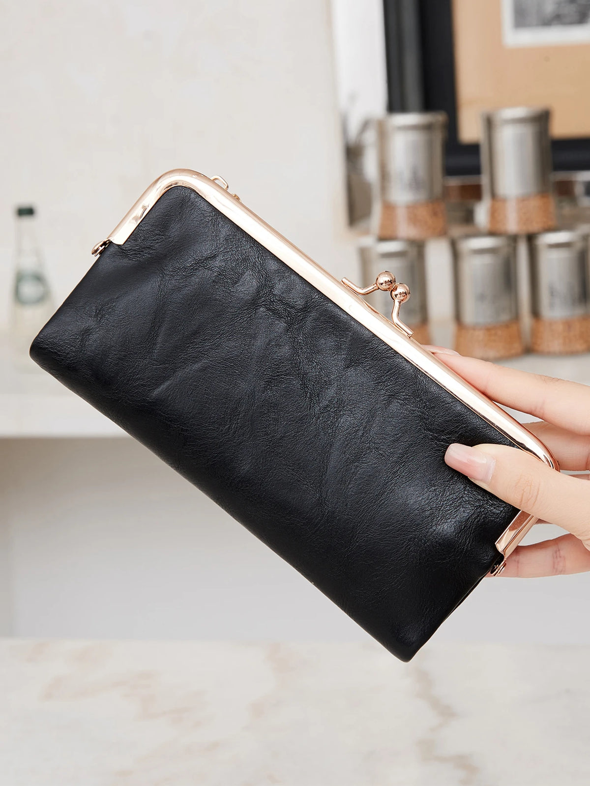 A black leather wallet with a gold-toned clasp is held by a hand against a light background.