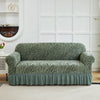A moss green jacquard sofa cover with an elastic skirt displayed on a white couch against a paneled wall.