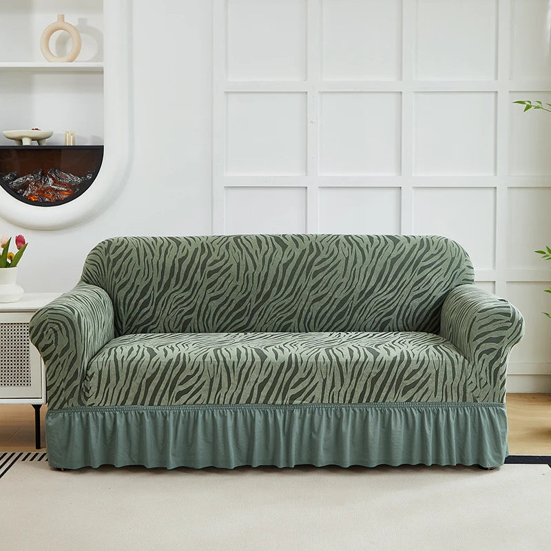 A moss green jacquard sofa cover with an elastic skirt displayed on a white couch against a paneled wall.