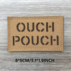 A rectangular tan woven patch with the text OUCH POUCH printed in dark lettering on a wooden surface.