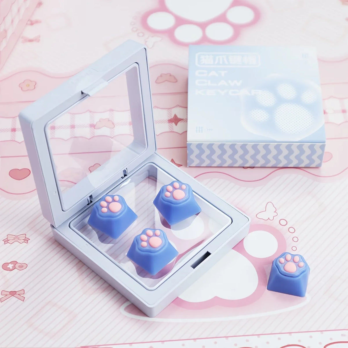 A clear plastic case holds silicone cat paw keycaps in blue and purple colors on a pink background.