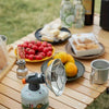 A portable butane gas stove is set up outdoors on a wooden table with various camping and outdoor items.