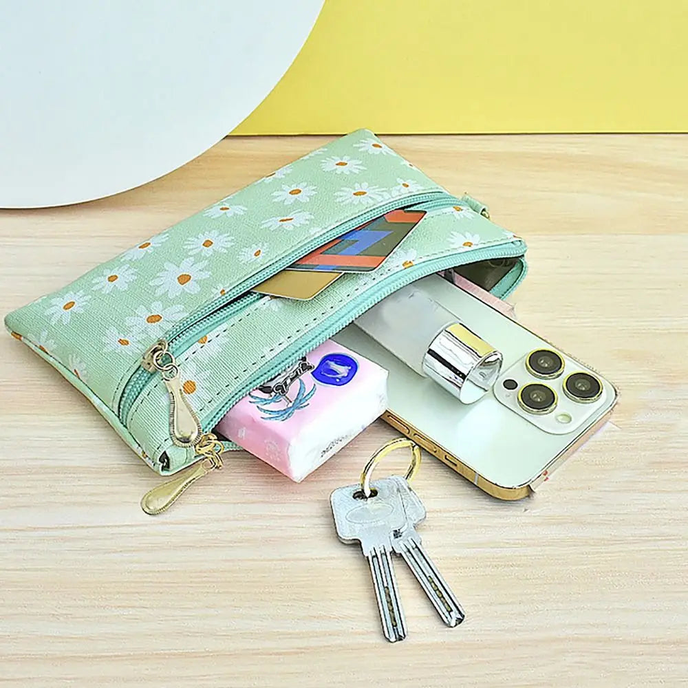A light green Daisy Print PU leather wristlet wallet with a zipper closure lying on a wooden surface next to a smartphone and