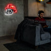 A man sits on a couch in a dimly lit room with a neon firefighter helmet sign glowing on the wall.