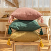 Three linen throw pillows in pink, green, and mustard yellow with tassels displayed on a wooden surface.