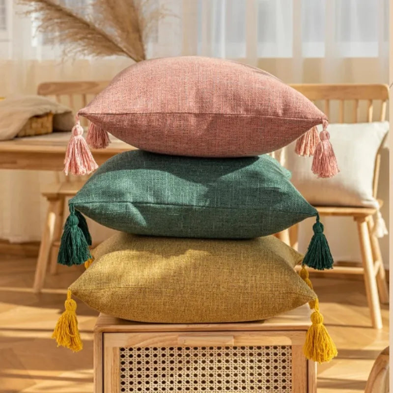 Three linen throw pillows in pink, green, and mustard yellow with tassels displayed on a wooden surface.