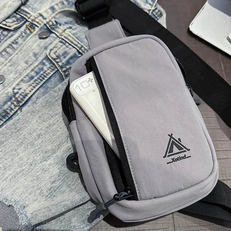 A gray water-resistant nylon chest sling bag with a black strap and a white interior lining is shown next to denim clothing.