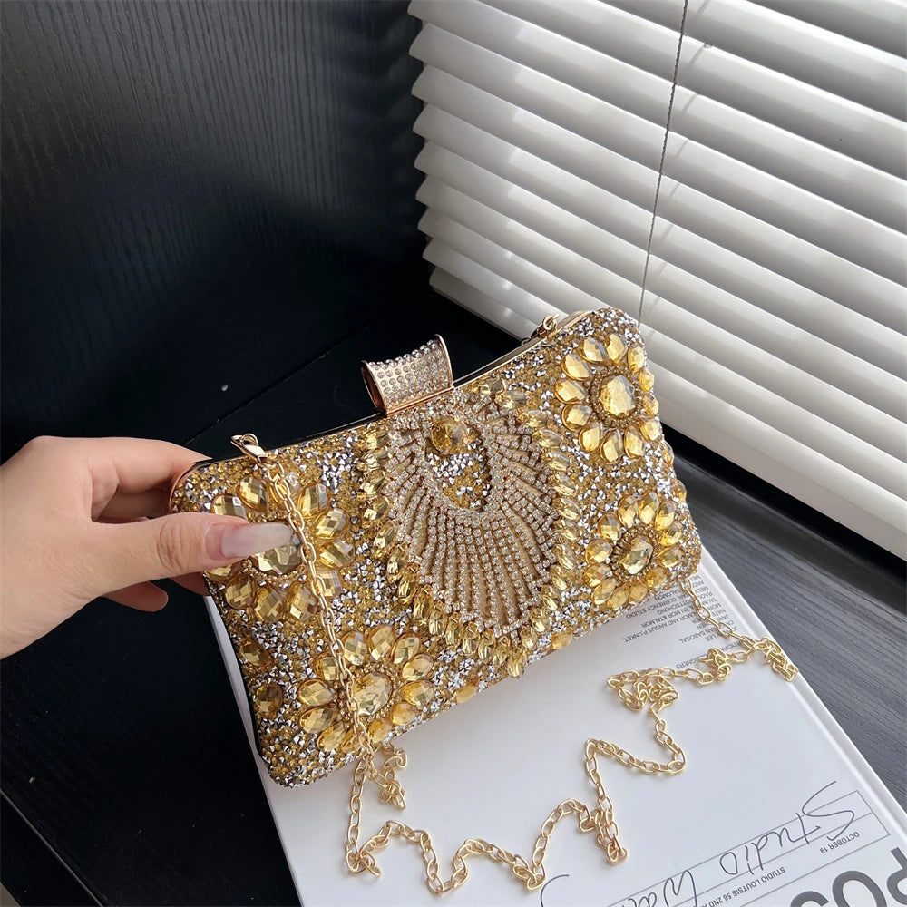 A gold rhinestone evening clutch with a crossbody strap is held against a dark background next to white blinds.