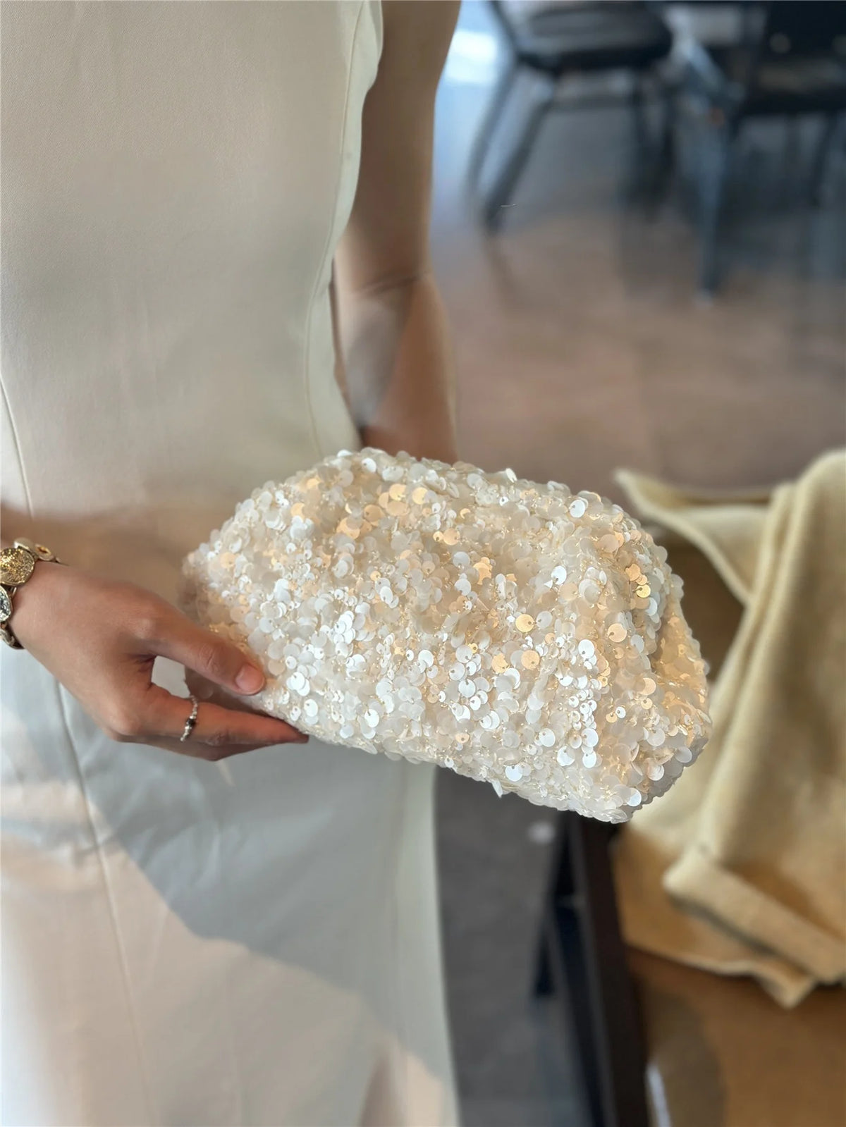 A hand holds a white sequin beaded chain shoulder clutch bag against a light-colored dress.