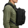A close-up of a person wearing a olive green shirt and a black crossbody chest bag with multiple zipper compartments and a MO