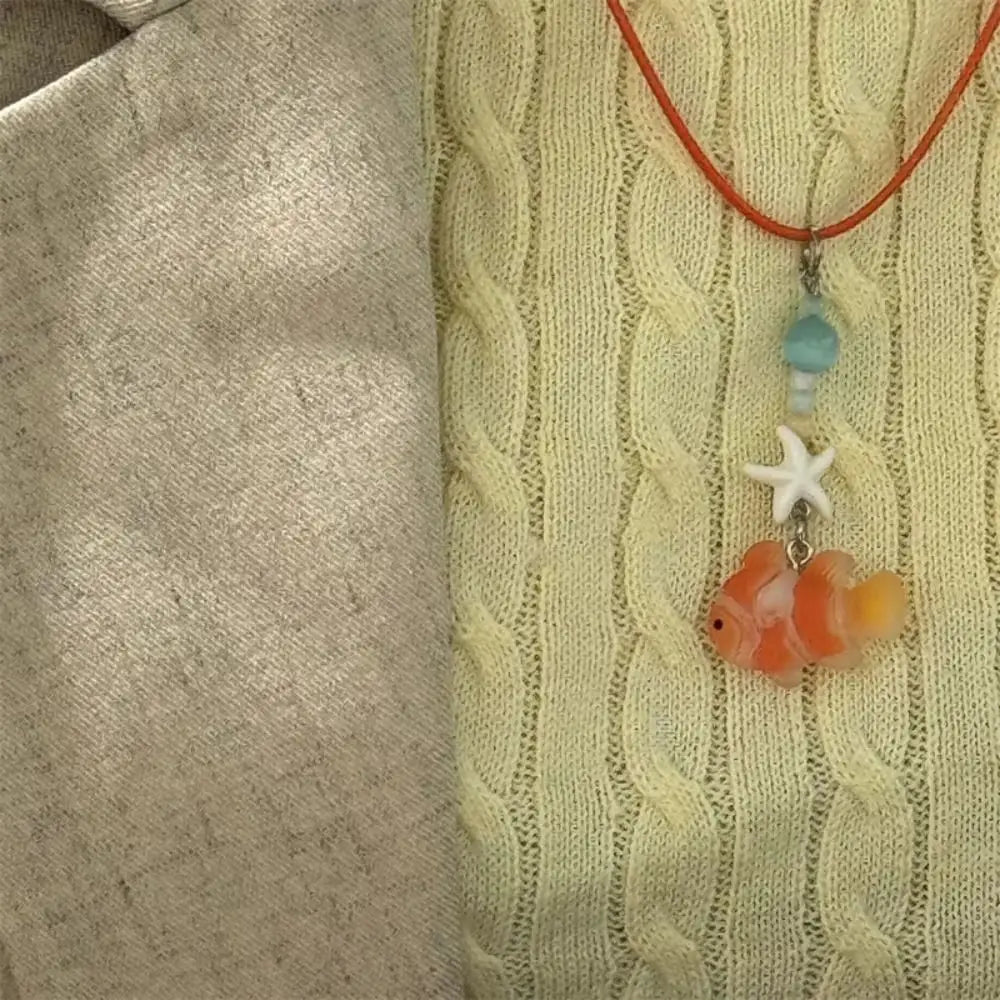 A necklace featuring a cute cartoon fish pendant with an orange body and white starfish charm on a yellow knitted sweater.
