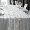 A white pearl beaded tulle table runner draped over a white tablecloth for an elegant wedding or event setting.