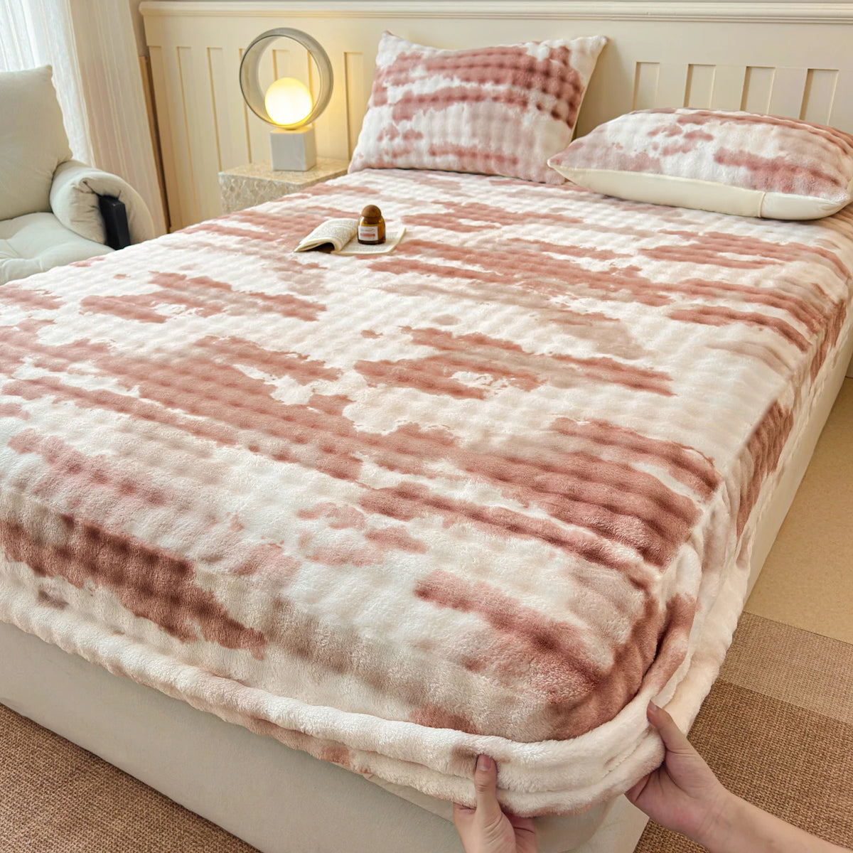 A cozy faux fur fitted sheet in pink and white stripes is displayed on a bed with pillows and a bedside lamp.