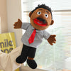 A soft plush full-body hand puppet resembling a man with dark skin, a mustache, and a red tie, standing in front of a window.