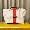 A white reusable elastic luggage strap with an orange-red strap and a wooden handle resting on a yellow surface.