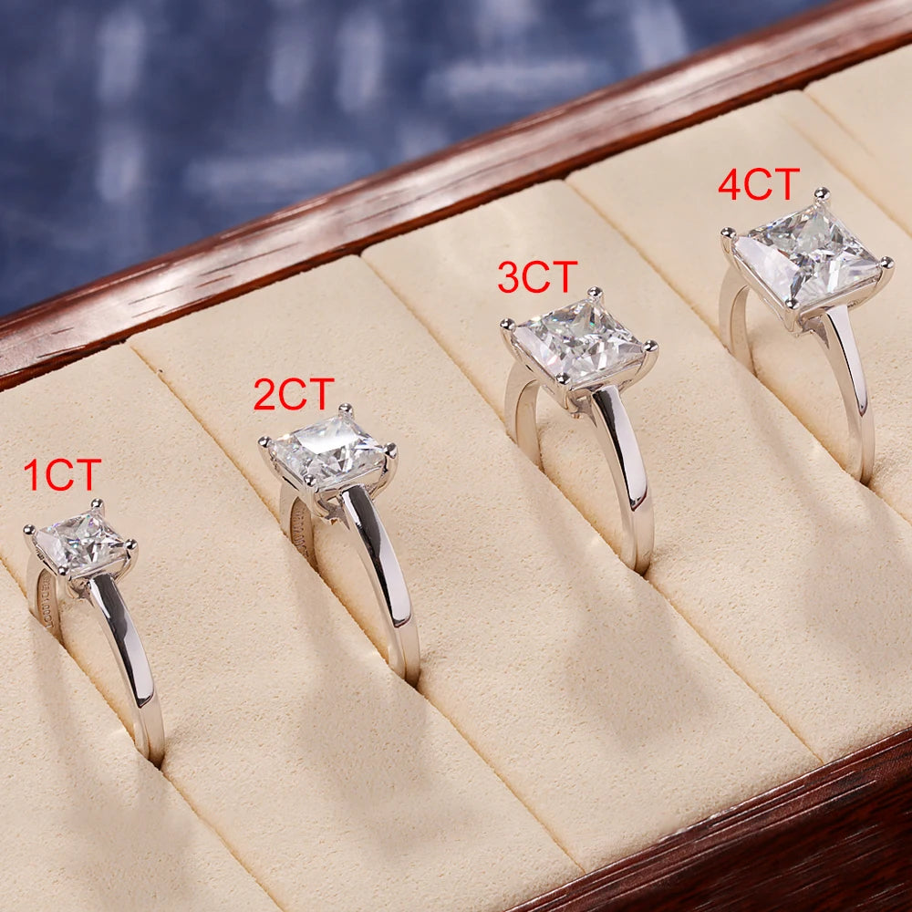 A display of four princess cut moissanite solitaire rings in 925 sterling silver arranged on a wooden tray.