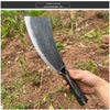 A hand holds a black forged steel axe with a curved blade in a dirt and plant environment.