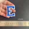 A hand holds a blue embroidered letter E patch, measured against a ruler.