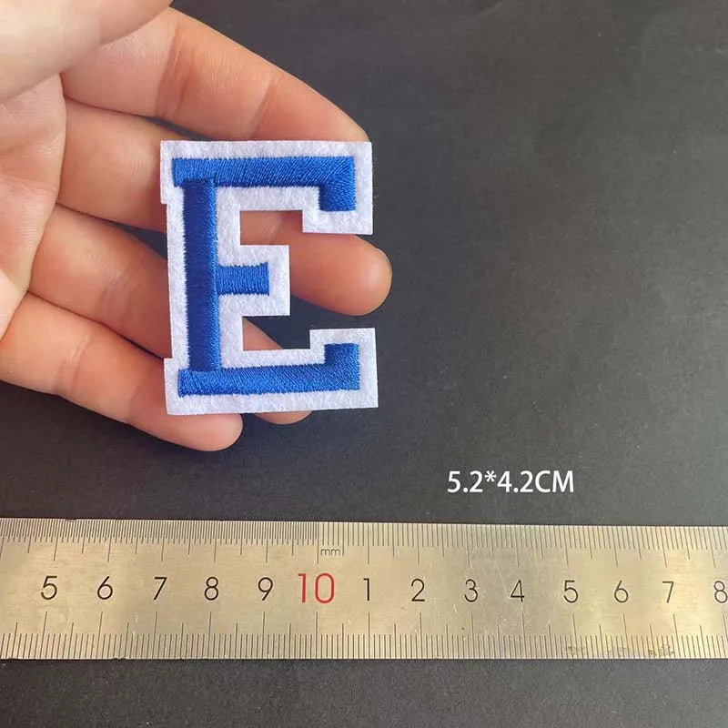 A hand holds a blue embroidered letter E patch, measured against a ruler.