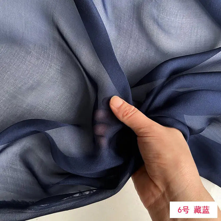 A hand is gently folding a piece of dark blue sheer silk chiffon fabric.