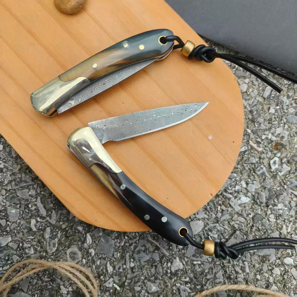 A folding knife with a Damascus steel blade and a water buffalo horn handle rests on a wooden surface.
