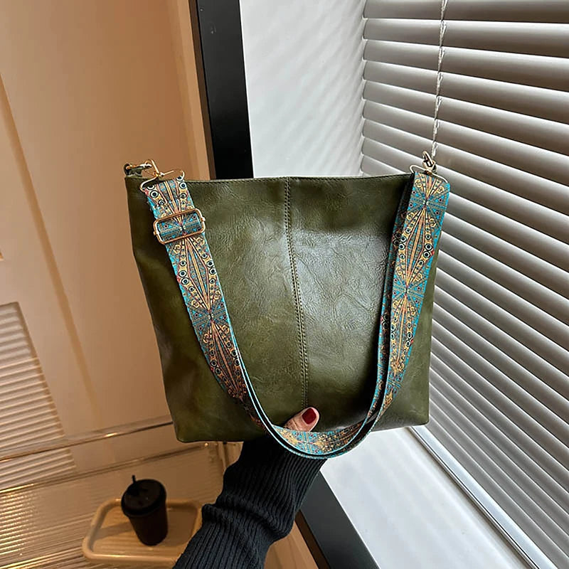Un sac à l'épaule vert olive foncé avec une sangle à motif, devant un store de fenêtre.