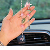 A hand holds a pink lotus car pendant featuring delicate pink flowers and a clear gemstone drop.