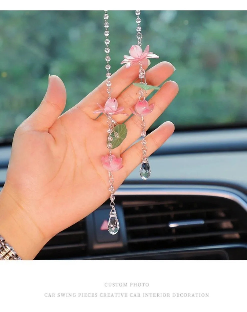 A hand holds a pink lotus car pendant featuring delicate pink flowers and a clear gemstone drop.