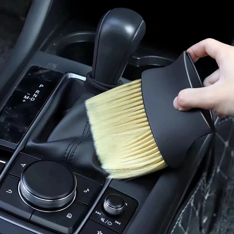 A hand is holding a black car interior detailing brush set with soft yellow bristles, positioned over a car gear shift.