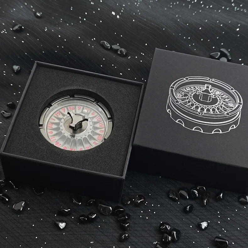 A black box containing a metal fingertip gyro roulette dice for tabletop games, resting on a dark surface with scattered blac